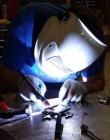 Swhift fabricator TIG welding a small stainless steel component behind a welding helmet